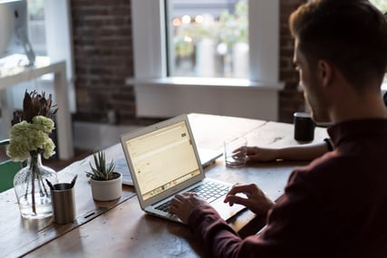 young man working on a laptop sitting at a wooden table with plants and vases on it and brick wall in background