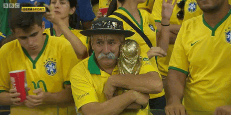 GIF sad Brazilian man in soccer outfit standing in crowd of fans hugging World Cup replica 
