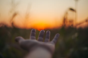 close up of hand reaching towards sunset horizon in a grass field